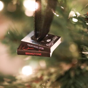 May include: A Christmas tree ornament shaped like two miniature books stacked together. The top book is titled "Quicksilver" and the bottom book is titled "Brimstone". The ornament is suspended by a black ribbon, with the books set against a backdrop of Christmas tree branches and lights.