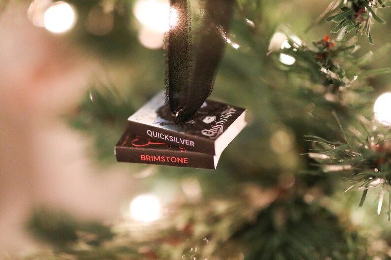 May include: A Christmas tree ornament shaped like two miniature books stacked together. The top book is titled "Quicksilver" and the bottom book is titled "Brimstone". The ornament is suspended by a black ribbon, with the books set against a backdrop of Christmas tree branches and lights.