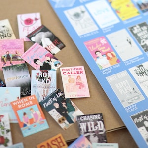 May include: A collection of miniature book covers with various titles and colorful designs, scattered on a brown surface. A blue sheet with more book covers is visible in the background. The books feature titles like "Happy Place" and "From Lukov with Love."