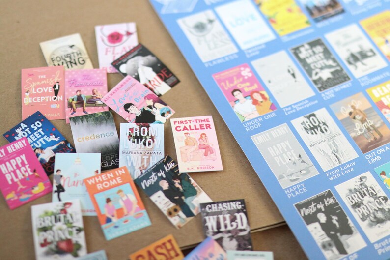 May include: A collection of miniature book covers with various titles and colorful designs, scattered on a brown surface. A blue sheet with more book covers is visible in the background. The books feature titles like "Happy Place" and "From Lukov with Love."