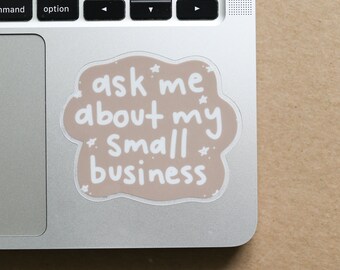 Puede incluir: Una pegatina con el texto "ask me about my Small business" en escritura blanca sobre un fondo en forma de nube marrón claro. Pequeñas estrellas blancas están dispersas alrededor del texto. La pegatina está sobre un portátil plateado.