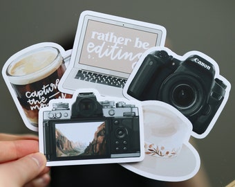 May include: A set of five stickers featuring a laptop, a coffee cup, two cameras, and a hat. The laptop has the text "rather be editing" on the screen. The coffee cup has the text "capture the moment" on the side. The cameras are black and have a vintage look. The hat is brown and has a white design.