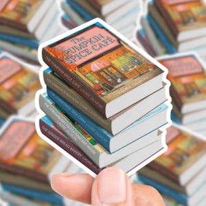 Puede incluir: Una pila de cinco pegatinas de libros en miniatura. La pegatina superior dice "The Pumpkin Spice Café" con una ilustración de una cafetería. Otros títulos incluyen "The Cinnamon Bun Book Store" y "The Christmas Tree Farm".