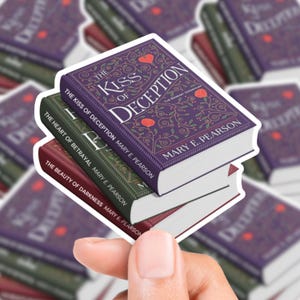 Puede incluir: Una pila de tres pegatinas de libros. El libro superior es morado y presenta el título "The Kiss of Deception". El libro del medio es verde y el libro inferior es burdeos. Todos los libros tienen bordes blancos.