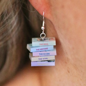 May include: Silver-toned dangle earring featuring a stack of miniature books. Each book has a different pastel-colored cover and a title printed on the spine: "The Deal", "The Mistake", "The Score", and "The Goal". The earring is attached to a silver fish hook.