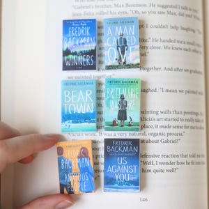 May include: A collection of miniature book replicas featuring titles by Fredrik Backman. The covers display various designs with titles like "Winners," "A Man Called Ove," "Bear Town," and "Us Against You." The books are held open on a page.