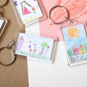 Peut inclure: Quatre porte-cl&eacute;s avec des cadres en plastique transparent affichant des dessins color&eacute;s d'enfants. Les dessins comprennent une fleur arc-en-ciel, une famille avec un ballon, une maison avec un soleil et une fille en robe.