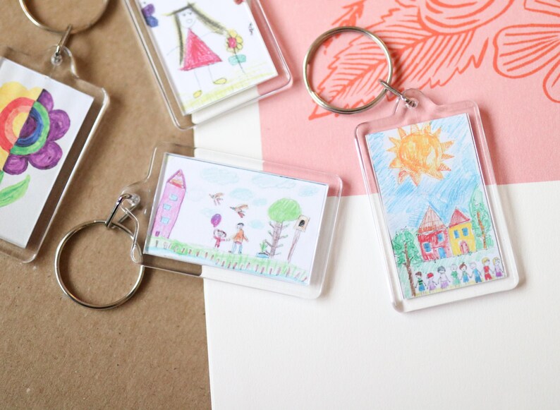 Peut inclure: Quatre porte-cl&eacute;s avec des cadres en plastique transparent affichant des dessins color&eacute;s d'enfants. Les dessins comprennent une fleur arc-en-ciel, une famille avec un ballon, une maison avec un soleil et une fille en robe.