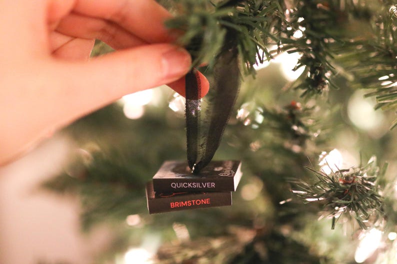 May include: A Christmas tree ornament with a black ribbon. The ornament is made of two small, rectangular blocks stacked on top of each other. The top block reads "QUICKSILVER" and the bottom block reads "BRIMSTONE" in red.