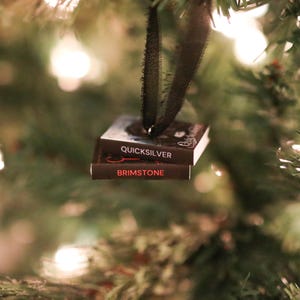 May include: A miniature book ornament with the titles "Quicksilver" and "Brimstone" in white and red lettering. The ornament hangs from a black ribbon against a blurred Christmas tree background with twinkling lights. A unique holiday decoration.