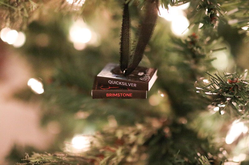 May include: A miniature book ornament with the titles "Quicksilver" and "Brimstone" in white and red lettering. The ornament hangs from a black ribbon against a blurred Christmas tree background with twinkling lights. A unique holiday decoration.