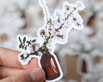 Puede incluir: Una pegatina que representa un jarrón marrón con flores blancas y tallos verdes. La pegatina tiene un borde blanco. Las flores tienen pequeños pétalos blancos. La pegatina está sujeta por una mano.