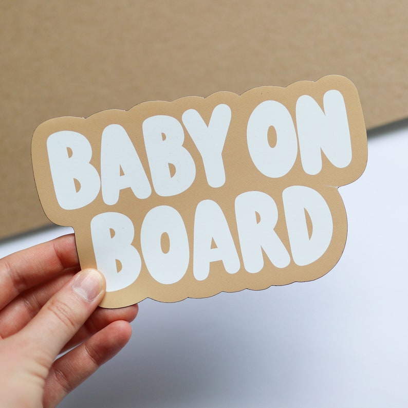 May include: A tan-colored car sign with white lettering that reads "BABY ON BOARD" in a rounded font.