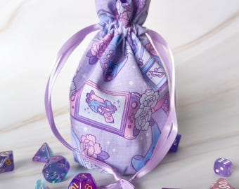 May include: A lavender drawstring bag with a repeating pattern of video game consoles, flowers, and stars. The bag is tied with a lavender ribbon and surrounded by various polyhedral dice in shades of purple, blue, and clear.