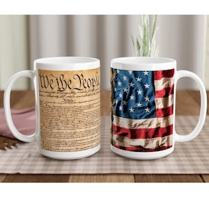 May include: Two white ceramic mugs. One mug features the text "We the People" in a vintage style, and the other displays a detailed American flag design. Both mugs have a white handle and a white interior. The mugs are on a checkered tablecloth.