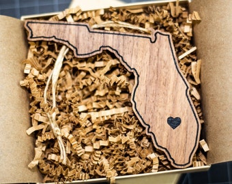 May include: A wooden cutout of the state of Florida with a small heart in the center. The cutout is in a brown box with brown packing paper.