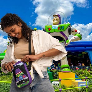 Puede incluir: Una mujer con una camisa blanca y pantalones vaqueros azules lleva un bolso bandolera morado y verde con una imagen de dibujos animados de Buzz Lightyear de la franquicia de pel&iacute;culas Toy Story. El bolso tiene una correa negra y un cierre de cremallera. La mujer est&aacute; de pie frente a una gran estatua de Buzz Lightyear.