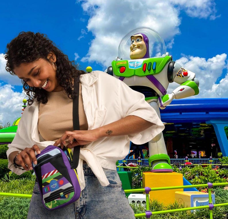 Puede incluir: Una mujer con una camisa blanca y pantalones vaqueros azules lleva un bolso bandolera morado y verde con una imagen de dibujos animados de Buzz Lightyear de la franquicia de pel&iacute;culas Toy Story. El bolso tiene una correa negra y un cierre de cremallera. La mujer est&aacute; de pie frente a una gran estatua de Buzz Lightyear.