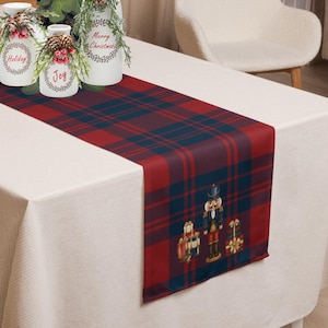May include: A red and navy blue plaid table runner with a Christmas theme. The runner features a nutcracker and gift box design at the bottom. The runner is placed on a white tablecloth.