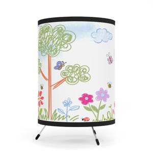May include: A white cylindrical lamp with a black top and bottom trim, supported by three metal legs. The lamp features a colorful, whimsical illustration of a tree, flowers, butterflies, bees, and a ladybug, set against a blue sky with a cloud.