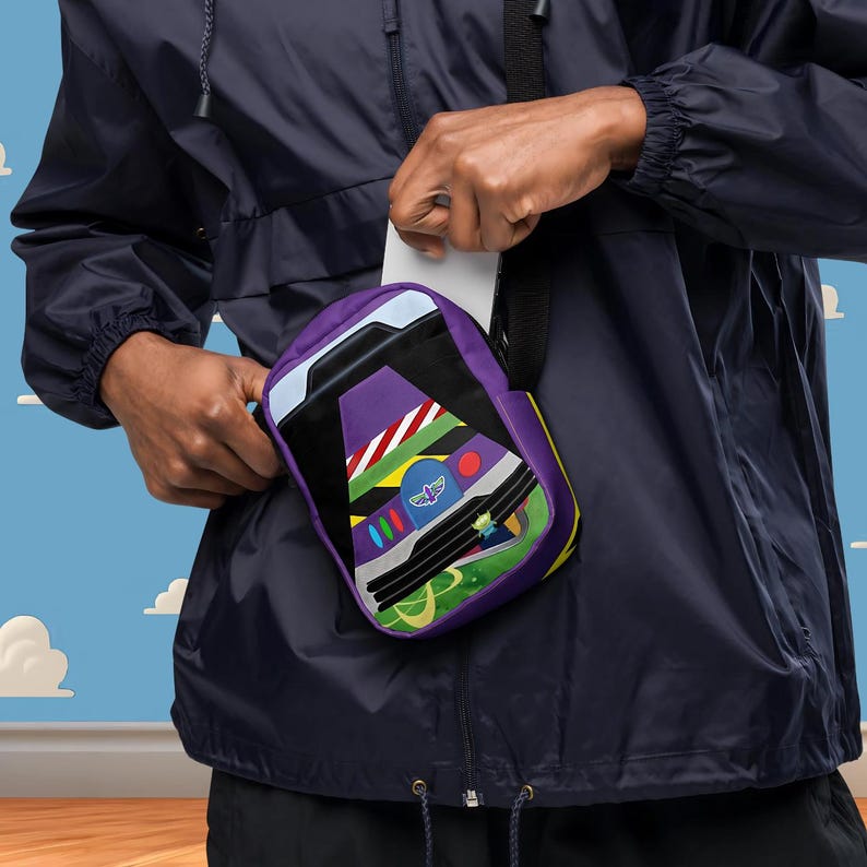 Puede incluir: Una bolsa de bandolera morada y verde con una imagen de dibujos animados de la nave espacial de Buzz Lightyear. La bolsa tiene una correa negra y un cierre de cremallera.