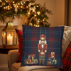 Puede incluir: Cojín decorativo cuadrado con un estampado de cuadros azul marino, rojo y verde. El cojín presenta una ilustración festiva de un soldado cascanueces y cajas de regalo envueltas. El cojín está colocado en un sofá beige.