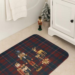 May include: A rectangular bath mat with a red and navy plaid pattern. The mat features a festive design with a nutcracker, a reindeer, and wrapped gifts. A white towel hangs over the edge of a white bathtub.