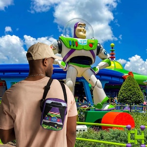 Puede incluir: Una persona lleva una gorra de b&eacute;isbol beige y una camisa rosa con una bolsa de bandolera morada y verde que presenta una imagen de dibujos animados de Buzz Lightyear de la franquicia Toy Story. La bolsa se lleva sobre el hombro izquierdo de la persona. La persona est&aacute; de pie frente a una gran estatua de Buzz Lightyear en un parque de atracciones.