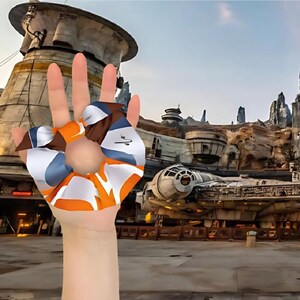 Puede incluir: Una mano sosteniendo una scrunchie azul, naranja y marrón con una franja blanca. La scrunchie tiene forma de lazo. El fondo es una imagen borrosa de un parque de atracciones temático de Star Wars.