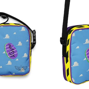 Puede incluir: Una bolsa de hombro azul y amarilla con una imagen de dibujos animados de una nave espacial de juguete p&uacute;rpura y blanca y la palabra "ANDY" en ella. La bolsa tiene una correa ajustable negra.