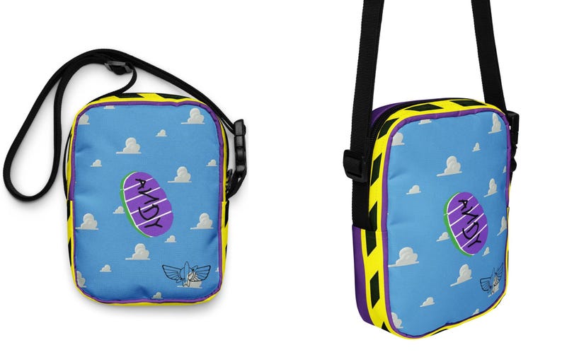 Puede incluir: Una bolsa de hombro azul y amarilla con una imagen de dibujos animados de una nave espacial de juguete p&uacute;rpura y blanca y la palabra "ANDY" en ella. La bolsa tiene una correa ajustable negra.