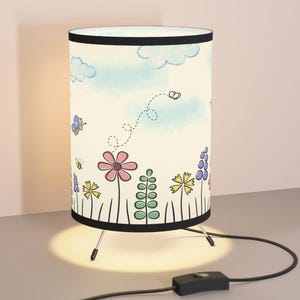 Puede incluir: Una lámpara de mesa blanca con una base negra y una pantalla cilíndrica. La pantalla presenta un diseño caprichoso dibujado a mano de flores, mariposas y abejas sobre un fondo de acuarela azul claro.