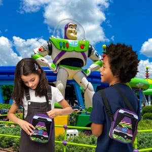 Puede incluir: Dos ni&ntilde;os llevan bolsas de bandolera moradas y negras con una imagen de dibujos animados de Buzz Lightyear de la pel&iacute;cula Toy Story. Las bolsas tienen un dise&ntilde;o verde, amarillo y blanco. Los ni&ntilde;os est&aacute;n de pie frente a una gran estatua de Buzz Lightyear.