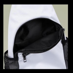 Puede incluir: Una mochila bandolera blanca con un compartimento frontal grande y abierto con forro interior negro. La bolsa incluye un cierre de cremallera negro y bolsillos laterales de malla negra a ambos lados, mostrando el espacio de almacenamiento.