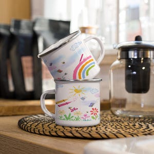 Puede incluir: Dos tazas de esmalte blanco con dibujos coloridos. La taza de arriba tiene un arcoíris y nubes, mientras que la taza de abajo tiene un sol, flores y lápices de colores. Las tazas están apiladas una encima de la otra.