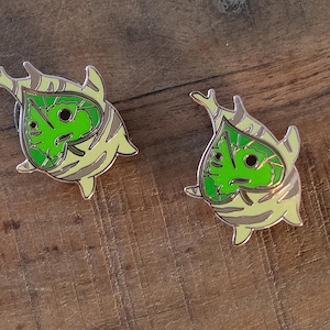 May include: Two enamel pins with a green leaf face design, set against a light yellow and grey background. The pins have a gold outline and are on a wooden surface.