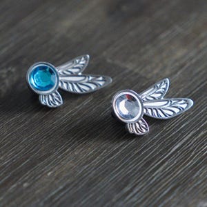 Könnte beinhalten: Zwei silberfarbene Libellenbroschen mit Edelsteinzentren. Eine Brosche hat einen blauen Edelstein, die andere einen klaren Edelstein. Die Broschen haben detaillierte Flügeldesigns auf Holz.