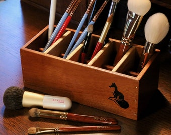 May include: A wooden makeup brush holder with six compartments, featuring a fox silhouette design. The holder is filled with various makeup brushes, including a large powder brush and a smaller angled brush. The holder is sitting on a wooden table.
