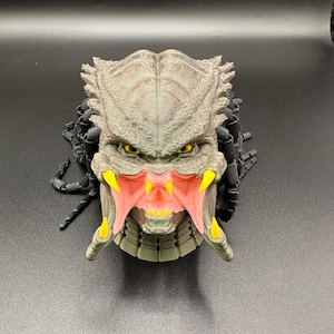 May include: A detailed 3D printed model of a Predator head with a textured gray skin, yellow eyes, and a red and yellow open mouth with sharp teeth. The head is surrounded by black dreadlocks.