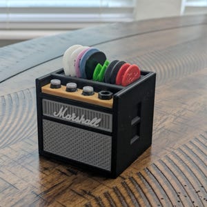 May include: A black and gray miniature Marshall amplifier with a wood top and a speaker grill. The amplifier has a slot for holding guitar picks, which are displayed in various colors.