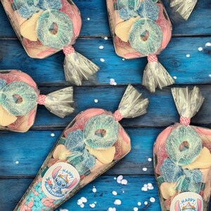 May include: Several clear cellophane bags filled with colorful candies, including blue, pink, and yellow sweets. Each bag is tied with a pink and white ribbon and features a sticker with a cartoon character and the words "Happy Birthday".