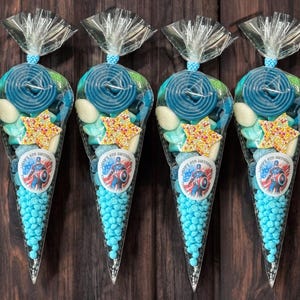 May include: Four transparent cone-shaped bags filled with blue sweets, spiral lollipops, star-shaped sprinkles, and white sweets. Each bag has a sticker with a superhero design and the text "Happy 4th Birthday". The bags are tied with a clear ribbon.