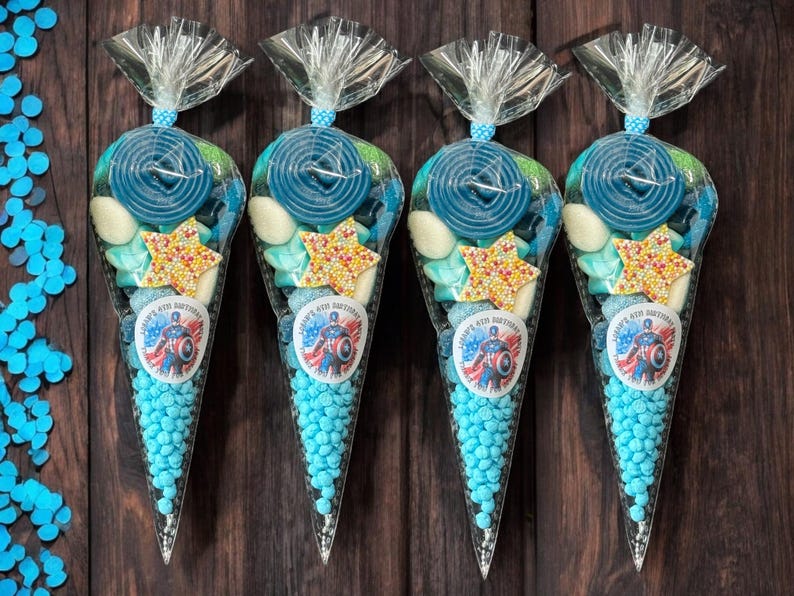 May include: Four transparent cone-shaped bags filled with blue sweets, spiral lollipops, star-shaped sprinkles, and white sweets. Each bag has a sticker with a superhero design and the text "Happy 4th Birthday". The bags are tied with a clear ribbon.