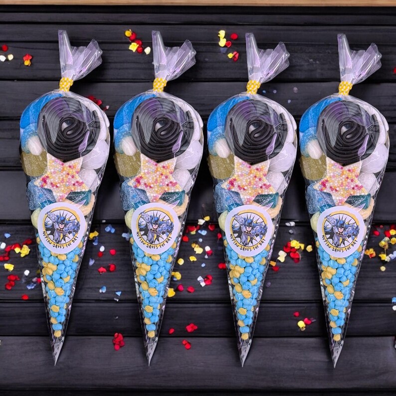 May include: Four transparent cone-shaped bags filled with sweets. Each bag has a Batman logo sticker and the text "Gotham Saves the Day". The sweets include blue and yellow candies, black licorice, and star-shaped sweets, tied with a gold ribbon.