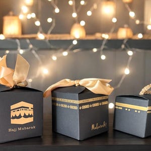 May include: Three black gift boxes with gold accents and gold ribbon. The boxes are decorated with Islamic designs and the text "Hajj Mubarak" and "Mubarak!" The boxes are sitting on a dark surface with a string of fairy lights in the background.