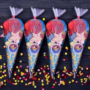 May include: Four cellophane cone bags filled with sweets. Each bag has a Wonder Woman logo sticker and is tied with a red and white polka dot ribbon. The bags contain red liquorice wheels, star-shaped sweets, and blue and yellow candies.