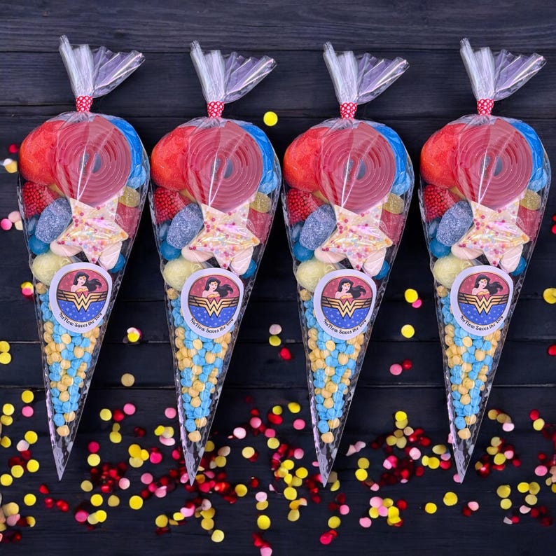 May include: Four cellophane cone bags filled with sweets. Each bag has a Wonder Woman logo sticker and is tied with a red and white polka dot ribbon. The bags contain red liquorice wheels, star-shaped sweets, and blue and yellow candies.