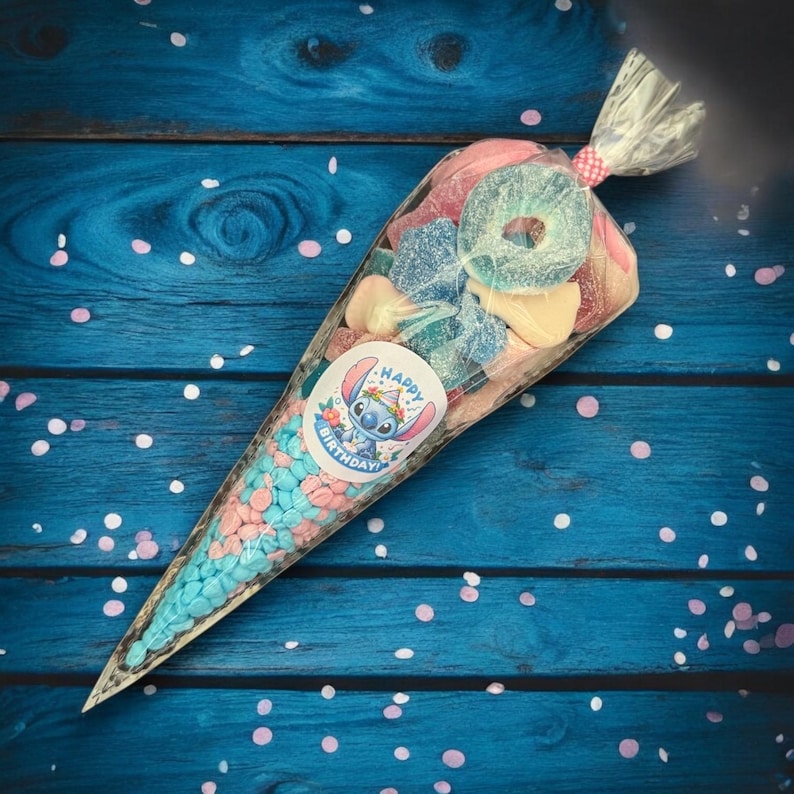 May include: A clear cellophane cone filled with colorful candies, including blue and pink sweets, and assorted gummy candies. A sticker with the character Stitch and the words "Happy Birthday" is visible on the cone. The cone is tied with a pink and white ribbon.