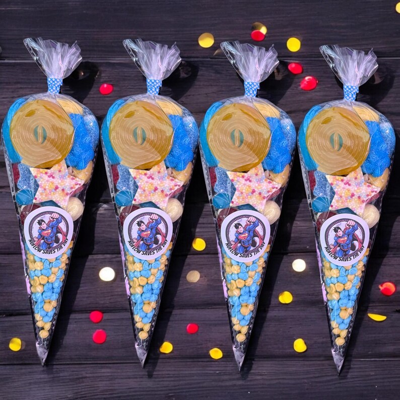 May include: Four cellophane cone bags filled with sweets. Each bag has a Superman logo sticker and is tied with a blue and white ribbon. The sweets include yellow and blue candies, star-shaped sweets, and a large yellow candy roll. The text on the sticker reads "SUPERMAN SAVES THE DAY".