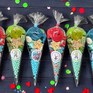 May include: Six cellophane cone-shaped bags filled with sweets. Each bag is tied at the top and features a different superhero sticker. The bags contain lollipops, star-shaped sprinkles, and other colourful treats. The bags are on a dark blue surface.